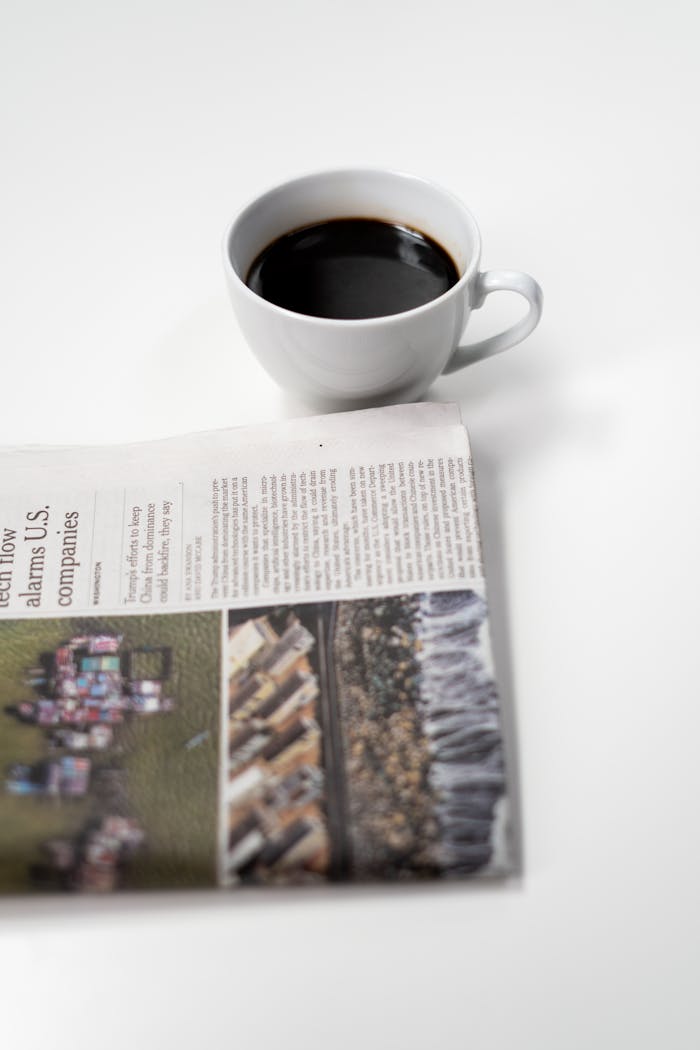 A white coffee cup with black coffee next to a folded newspaper on a bright table setting.