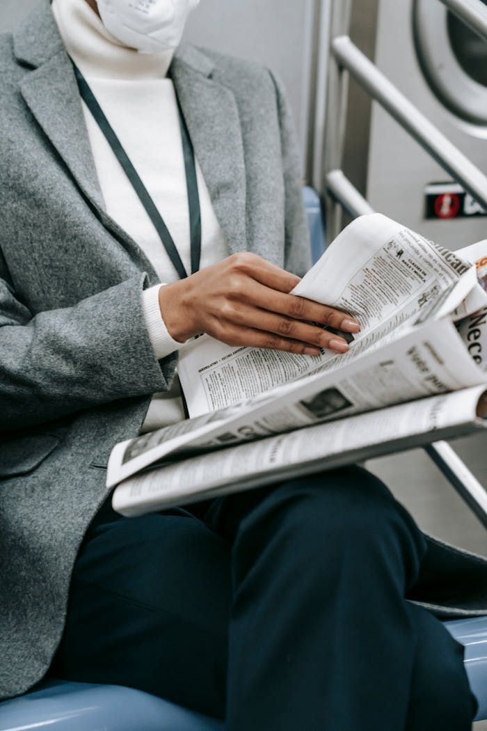 Crop anonymous black female entrepreneur in elegant outfit and medical mask reading newspaper while riding train